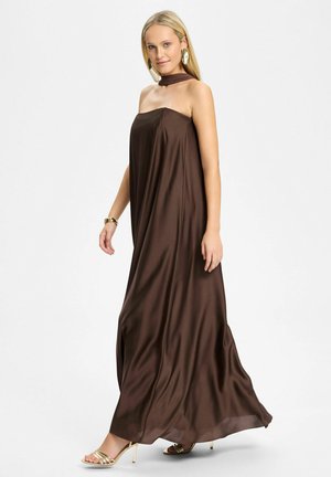 Donna con capelli biondi che indossa un abito lungo marrone senza spalline e fluente, sandali dorati con tacco alto, grandi orecchini dorati e un braccialetto dorato.