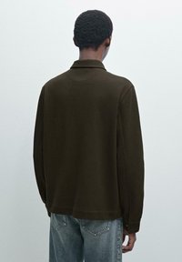 Brun langærmet polo skjorte, tekstureret stof, standard krave og knapærmer. Båret med slidte blå jeans, set fra ryggen.