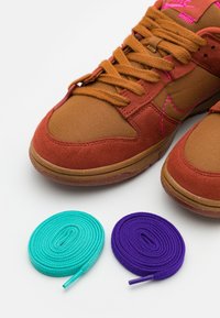 Sneaker marroni e ruggine con cuciture rosa accanto a due lacci da scarpa arricciati, uno turchese e uno viola, su uno sfondo bianco.