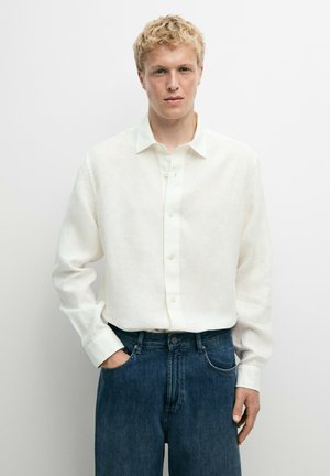 Ung mand med kort blondt hår iført en hvid langærmet skjorte og blå jeans, stående mod en ensfarvet hvid baggrund.