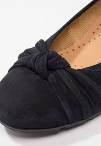 Gros plan sur une chaussure plate en daim noir avec un nœud en tissu torsadé à l'avant et une semelle intérieure beige perforée et rembourrée.