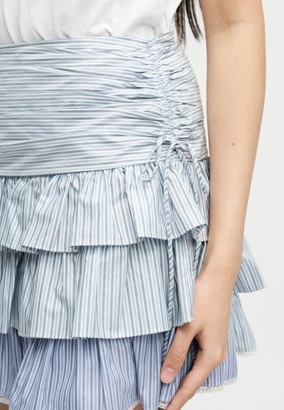 Λεπτομέρεια μέσης και φούστας που δείχνει ύφασμα με κατακόρυφες ρίγες σε μπλε και άσπρο, με πτυχώσεις και δέσιμο στο πλάι καθώς και στρωματωτές βολάν στο κάτω μέρος του κορμού ενός ατόμου.