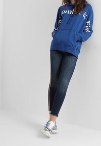 Blå hoodie med vit text, framficka, kombinerad med mörka jeans med sidostänger och fransade fållar, avslutad med färgglada sneakers.