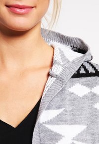 Grå stickad tröja med huva, som har ett geometriskt mönster i svart och vitt, framhävt av texturerad tyg och en slät V-ringning.