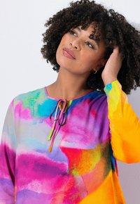 Vrouw met krullend haar die een kleurrijke, tie-dye patroon top draagt, haar haar aanraakt en omhoog kijkt tegen een effen lichte achtergrond.