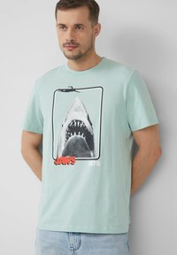 Světle modré bavlněné tričko s grafikou otevřené tlamy žraloka, červeným textem "JAWS" a "1975" pod ním, kulatý výstřih, krátké rukávy.