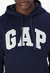 Sudadera con capucha de color azul marino con un gran logo "GAP" en letras bordadas de color gris; material de algodón, bolsillo tipo canguro.