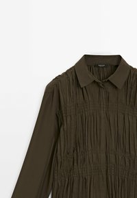 Chemise vert olive foncé à manches longues avec un col pointu et une texture plissée verticale sur le devant et le haut des bras.