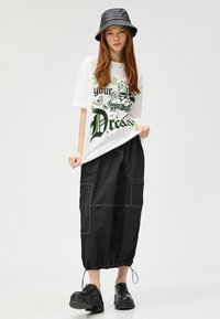 Camiseta gráfica blanca con estampado verde y negro, combinada con pantalones holgados negros con acentos de costura y puños ajustables. Zapatos de plataforma negros.