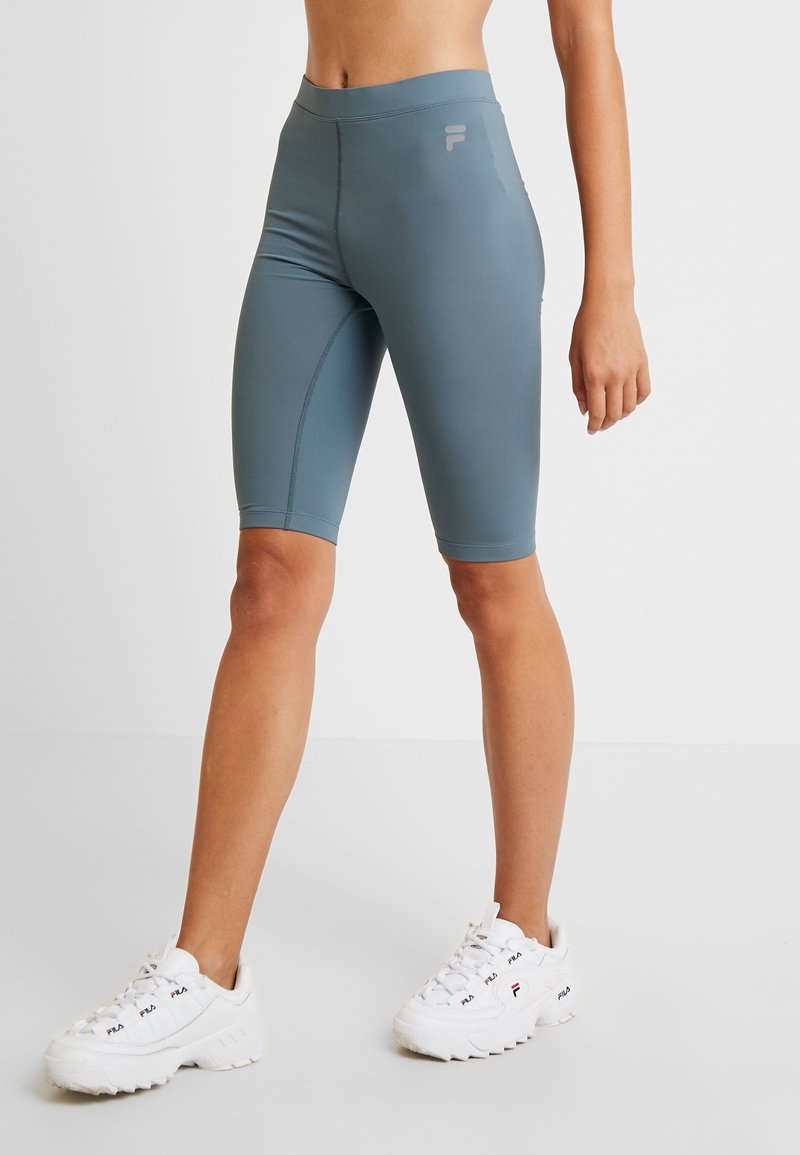 Femme portant un short cycliste bleu-gris taille haute et des baskets blanches FILA, debout sur un sol blanc sur fond uni.