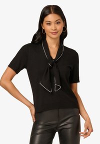 Schwarzer kurzärmeliger Pullover mit Knoten am Ausschnitt und kontrastierender weißer Naht. Weiches Strickmaterial, figurbetonte Passform, getragen mit schwarzen Hosen.
