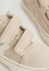 Beige Sneakers mit zwei Klettverschlüssen, aus Canvas-Material, runder Zehenform, strukturierten Akzenten und einer Gummisohle mit einem dezenten Muster.