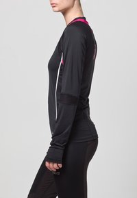Donna in una maglietta nera a maniche lunghe da allenamento con accenti rosa e leggings neri, in piedi di lato su uno sfondo grigio chiaro.
