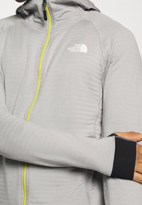 Chaqueta de cremallera gris claro hecha de un material texturizado, con una cremallera amarilla y acentos en los puños en negro. Logos mostrados en la zona del pecho.