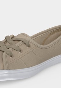 Μπεζ sneaker με κορδόνια, υφή στο επάνω μέρος, ραμμένα λεπτομερή στοιχεία, επενδεδυμένος γιακάς και λευκή λαστιχένια σόλα, εμφανίζεται σε κοντινή πλευρική λήψη.