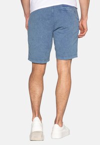 Siniset farkkushortsit, joissa on rento leikkaus, näkyvät ompeleet, takatasku ja sileä pinta, yhdistettyinä valkoisiin tennareihin.