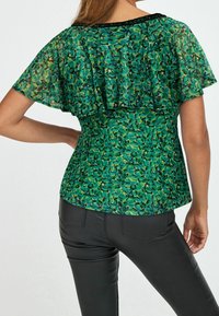 Blusa floreale verde con maniche trasparenti, corpino aderente e collo bordato di nero. Abbinata a pantaloni neri attillati.