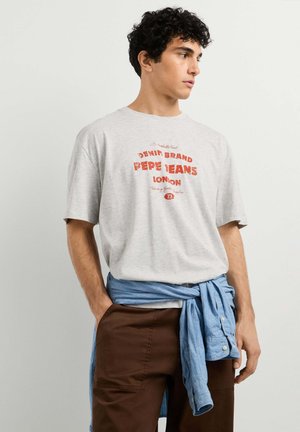 Giovane uomo con capelli ricci indossa una t-shirt Pepe Jeans London grigio chiaro, pantaloni marroni e una camicia blu legata in vita, guardando di lato.