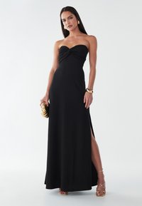 Melna bezplecu maksi kleita ar savīti augšdaļu, sānu šķēlumu un gludu tekstūru. Nēsāta ar zelta aksesuāriem un atvērta purngala sandales.