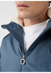 Chaqueta azul con cuello texturizado, forro interior blanco y cremallera visible con tirador circular. Primer plano del área del cuello.
