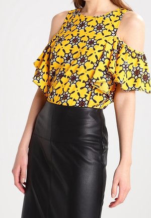 Kobieta w żółtej bluzce cold-shoulder z czarno-białymi wzorami gwiazd oraz czarnej wysokiej skórzanej spódnicy.