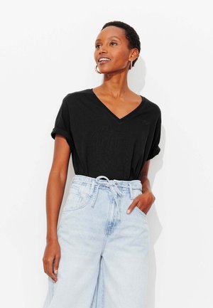 Femme portant un t-shirt noir à manches courtes avec col en V, rentré dans un jean taille haute large bleu clair à ceinture coulissante, debout avec une main dans la poche.