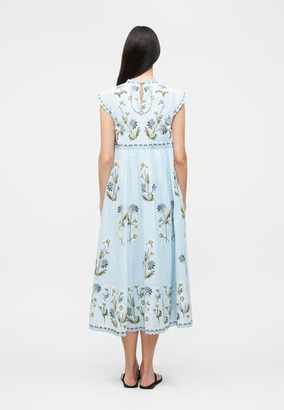 Γυναίκα με μακριά σκούρα μαλλιά που φοράει ανοιχτό μπλε αμάνικο φόρεμα με κεντήματα λουλουδιών, στέκεται με την πλάτη γυρισμένη, φορώντας μαύρα φλατ σανδάλια.