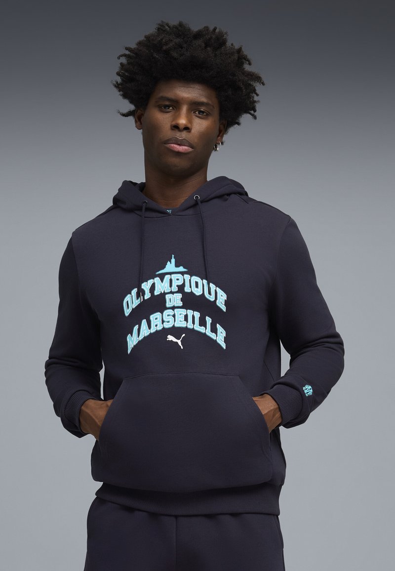 Sweat à capuche bleu marine avec graphique "Olympique de Marseille" en bleu clair. Comprend une poche frontale et des poignets côtelés. Fabriqué en tissu doux.