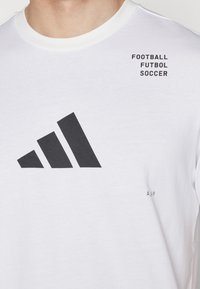 T-shirt bianca in cotone con logo a tre strisce nere e la scritta "FOOTBALL FUTBOL SOCCER" stampata sul petto. Testo in un carattere sans-serif in grassetto.
