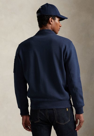 Homme portant une casquette de baseball bleu marine, un sweat bleu marine et un jean bleu foncé, debout avec le dos légèrement tourné contre un fond uni.