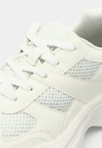 Λευκό αθλητικό παπούτσι με επάνω μέρος από δέρμα με υφή και πλέγμα, που διαθέτει επίπεδα κορδόνια, διακοσμητικό ράψιμο και μαλακή, στρογγυλεμένη σόλα.