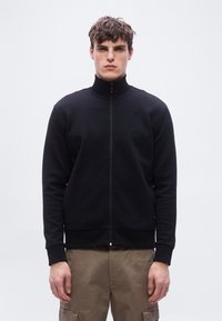 Νεαρός άνδρας που φοράει μαύρο μπουφάν με φερμουάρ και ψηλό γιακά, καθώς και καφέ cargo παντελόνι, στέκεται μπροστά σε απλό φωτεινό φόντο.