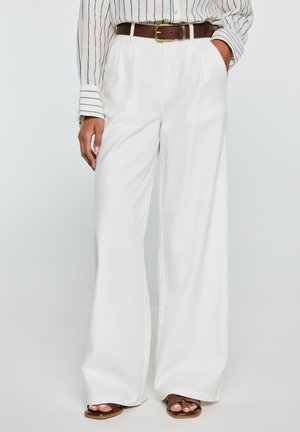 Kobieta ubrana w białe spodnie o szerokich nogawkach, brązowy pasek, pasiastą bluzkę z długim rękawem i brązowe sandały na otwartym palcu, stojąca z jedną ręką w kieszeni.
