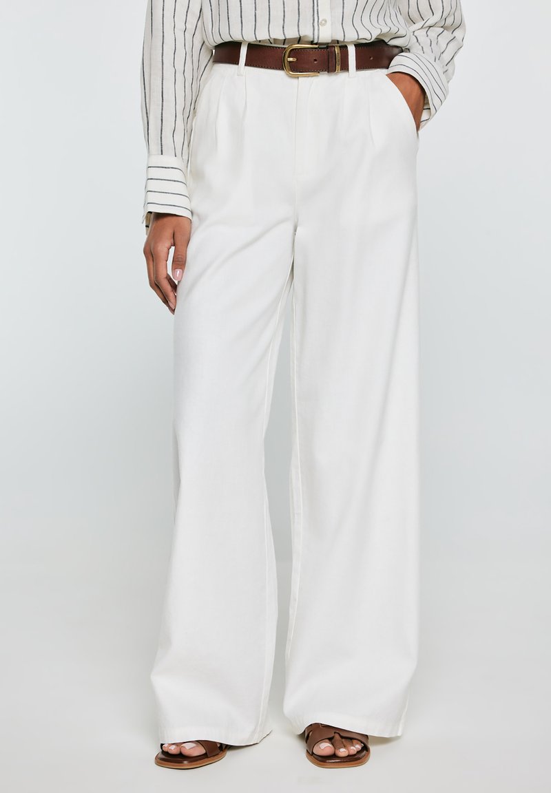 Žena oblečená v bílých kalhotách s širokými nohavicemi, hnědým opaskem, pruhovaném tričku s dlouhým rukávem a hnědých sandálech s otevřenou špičkou, stojící s jednou rukou v kapse.