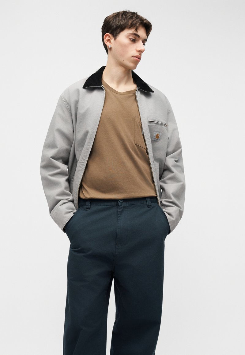 Junger Mann trägt graue Jacke mit Reißverschluss und schwarzem Kragen, braunes T-Shirt und dunkelblaue Hose, Hände in den Taschen, schaut seitlich nach unten.