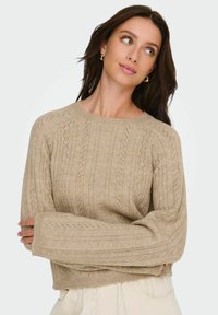 Pull beige en tricot avec texture côtelée, doté d'un col rond et de manches longues avec des motifs détaillés sur le tissu.