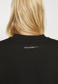 Svart sweatshirt med ribbad krage och en liten vit broderad logotyp på ryggen, med en slät textur och en åtsittande form.