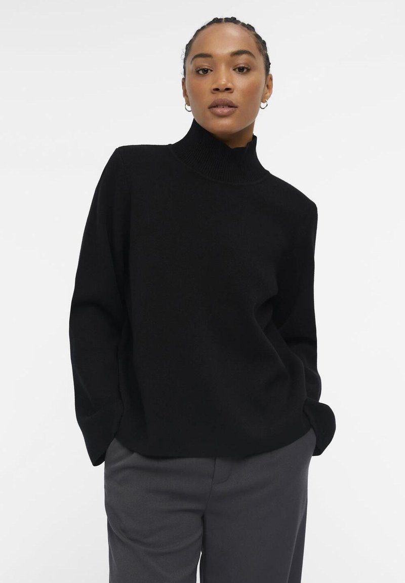 Maglione nero a collo alto con colletto a coste, vestibilità ampia, maniche lunghe e tessuto morbido, abbinato a pantaloni grigi.