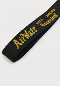 Μαύρο υφαντό λουρί από ύφασμα με κίτρινα γράμματα που αναγράφουν «AirWair with Soles Bouncing». Η υφή είναι ριμπ, προσφέροντας κράτημα και αντοχή.