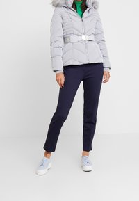 Femme portant une doudoune grise claire avec capuche en fourrure et ceinture, haut vert, pantalon bleu marine, et baskets compensées bleu clair sur fond blanc.