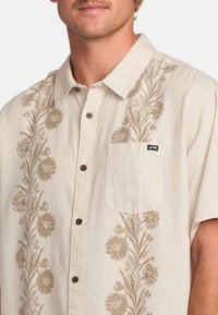 Camisa de manga corta beige claro con un patrón floral marrón, que presenta un diseño de botones y un bolsillo en el pecho con una etiqueta de la marca.