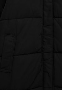 Veste matelassée noire avec une texture lisse, présentant des sections matelassées et une poche latérale. Le design est minimaliste sans quincaillerie visible.