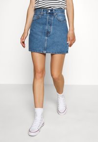 Kvinna som bär en blå denimminikjol, randig topp, vita strumpor och vita högsnörade sneakers som går mot en enkel vit bakgrund.