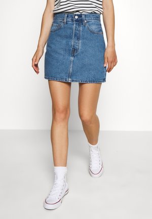 Donna che indossa una minigonna di jeans blu, una maglietta a righe, calze bianche e sneakers alte bianche mentre cammina su uno sfondo bianco uniforme.
