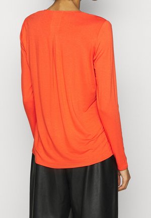 Persoon draagt een feloranje longsleeve met een centrale rugplooi, gecombineerd met losse zwarte broek, gezien van achteren.