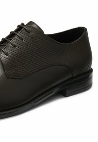 Scarpa in pelle marrone scuro con finitura liscia e parte superiore perforata. Suola robusta con accenti neri e logo vicino al tallone.
