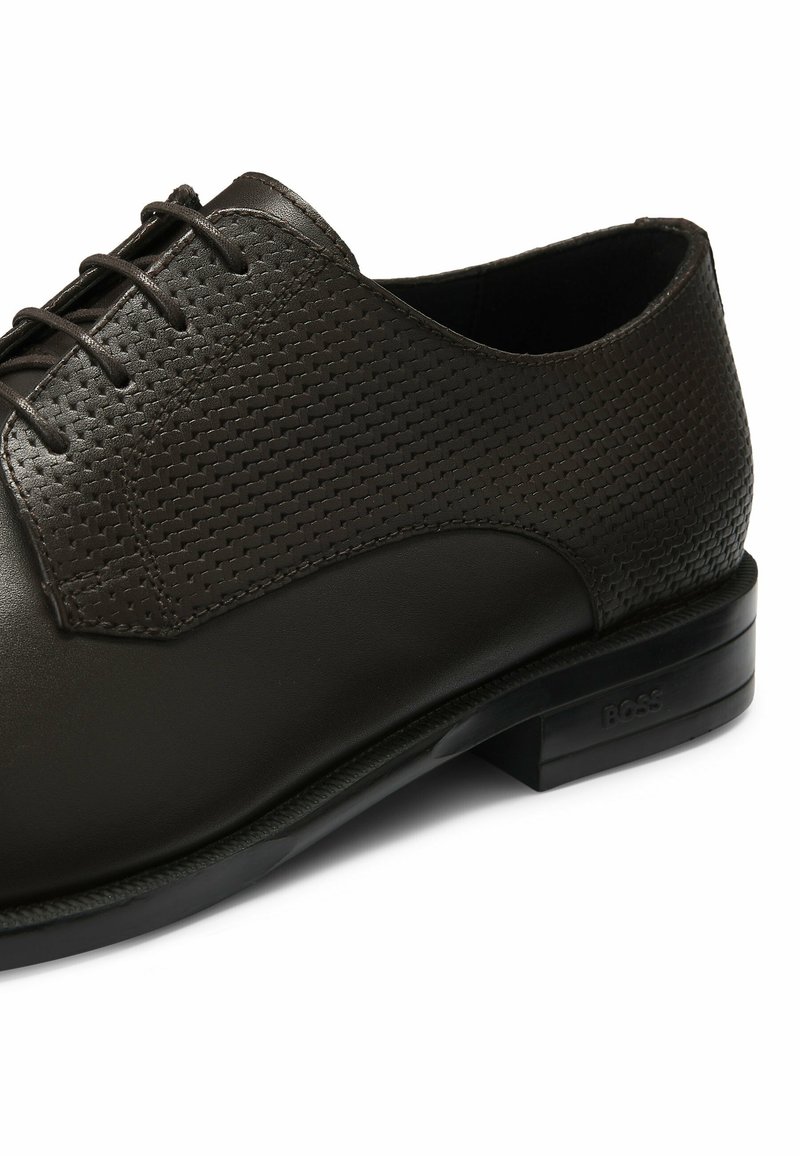 Chaussure en cuir marron foncé avec une finition lisse et une section supérieure perforée. Semelle robuste avec des accents noirs et un logo près du talon.