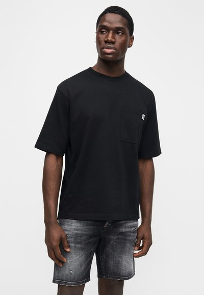 Črna majica s kratkimi rokavi iz bombaža, z žepom na prsih in majhnim logotipom. Nosena z raztrganimi sivo modrimi kratkimi hlačami.