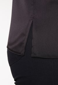 Black blouse made of smooth fabric, featuring a side slit hem. Paired with fitted black pants, showing a close-up view of the transition.