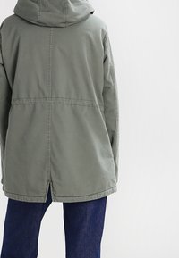 Olivegrüner Kapuzenjacke mit lockerer Passform, gerade geschnittenem Saum und sichtbaren Nahtdetails, getragen über blauen Jeans.